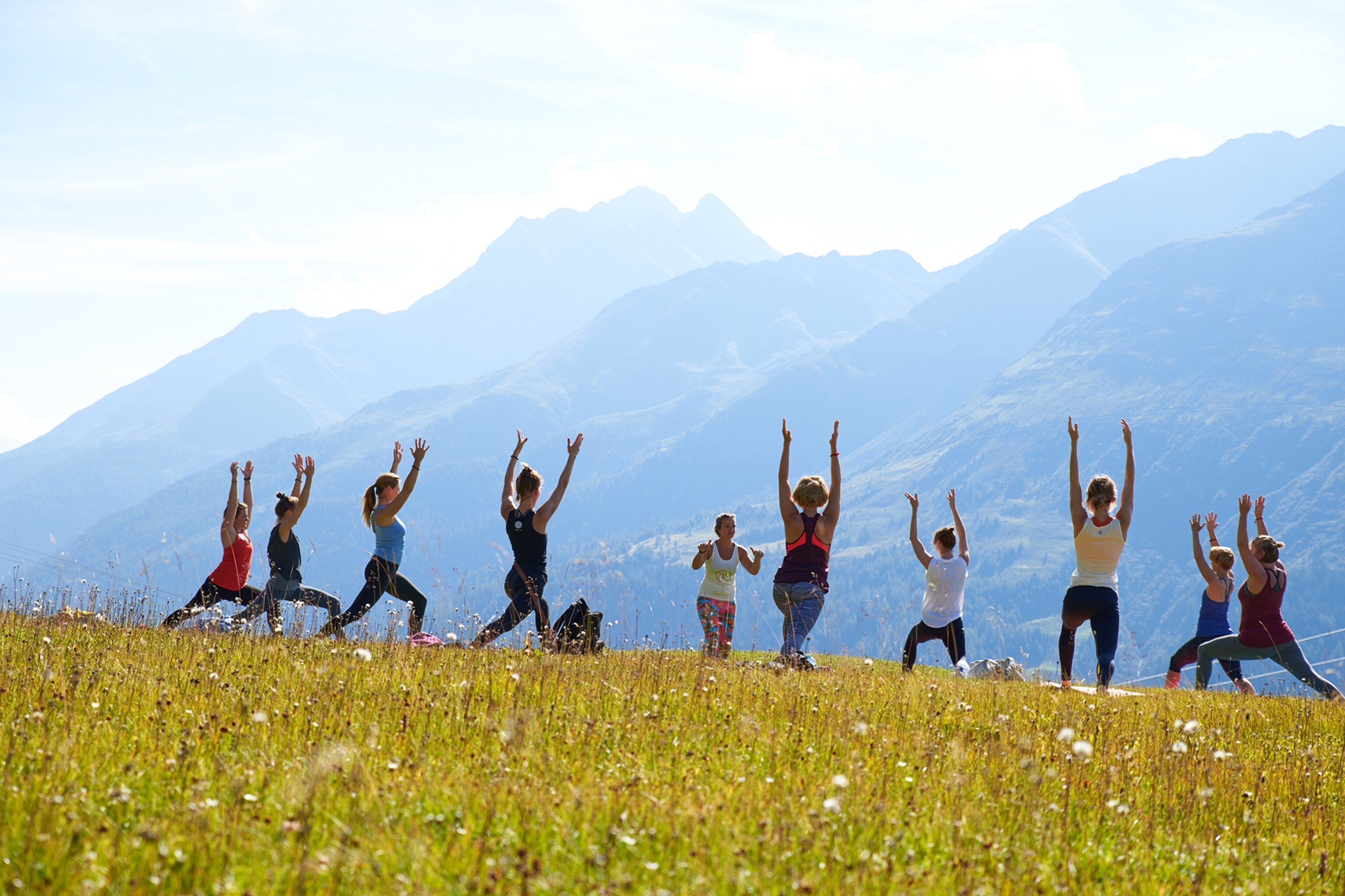 People doing yoga on a grassy hill with mountains in the background.