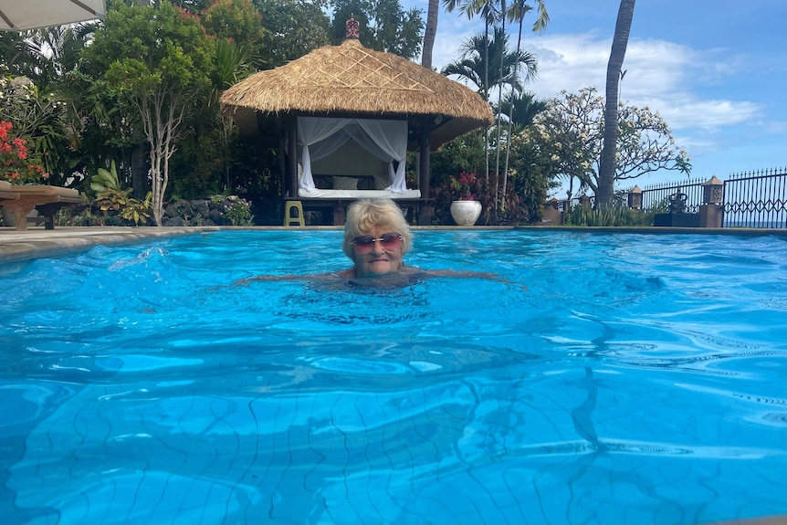 A woman with short grey-blond hair and sunglasses on swims in a blue pool with a thatch-roofed gazebo next to it.