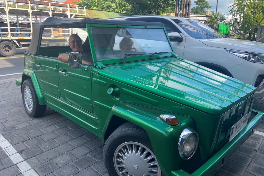 A man and woman sit in a small green 1971 Safari car with a soft top. The man has his elbow on the door and smiles happily. 