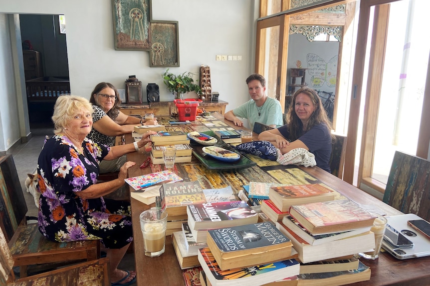 Three women and one man sit around a large table covered in books and a tray with pieces of cake.