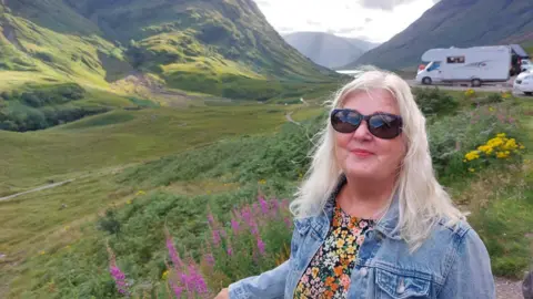 Supplied Nicky Maidment has medium length bleach blonde hair and wears a floral top, denim jacket and sunglasses.
She stands in a meadow near some mountains.