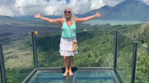 Supplied Nicky Maidment stands with her arms outstretched on a glass balcony overlooking some mountains. She wears a light blue tank top and a white frilly skirt.