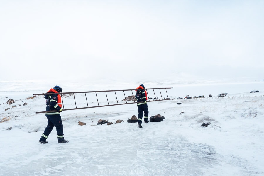 Two EMS workers dressed in life jackets carry a long ladder across an icy road to the shore of Paravani Lake.