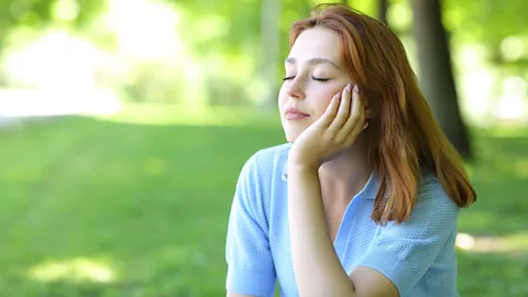 Alamy A woman sits with her eyes closed in a sunny, green space (Credit: Alamy)