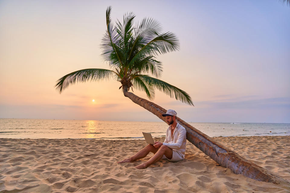 Young nomad man freelancer sitting under a palm tree and working remotely online at a laptop, enjoying a dream job anywhere in the world