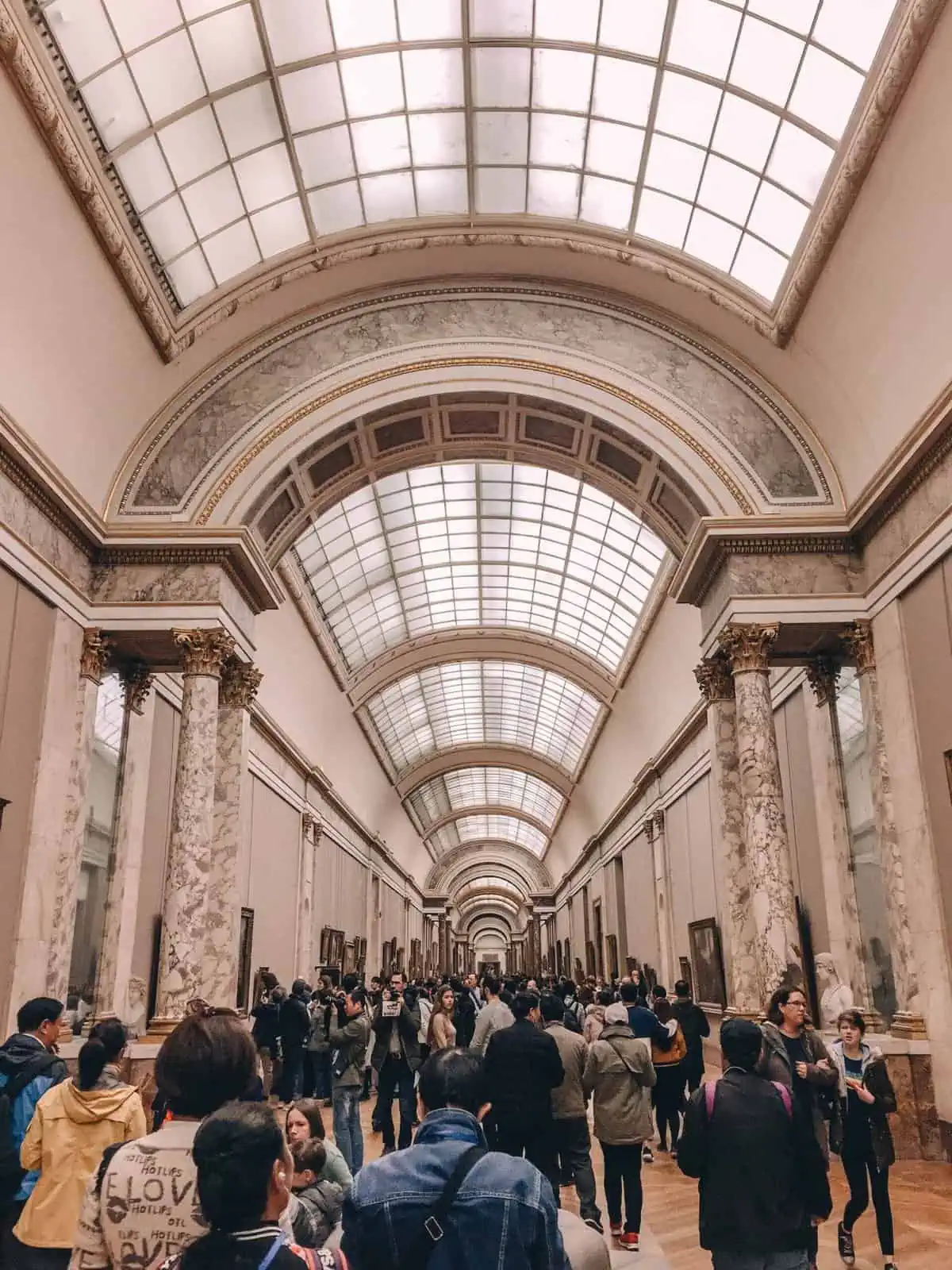 A long hallway in the Louvre filled with art on the walls and people walking down the corridor 