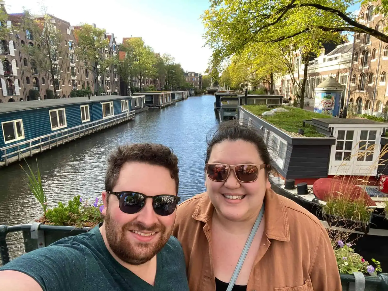 Riana and Colin taking a selfie on the canals in amsterdam with trees, houseboats and skinny houses behind them