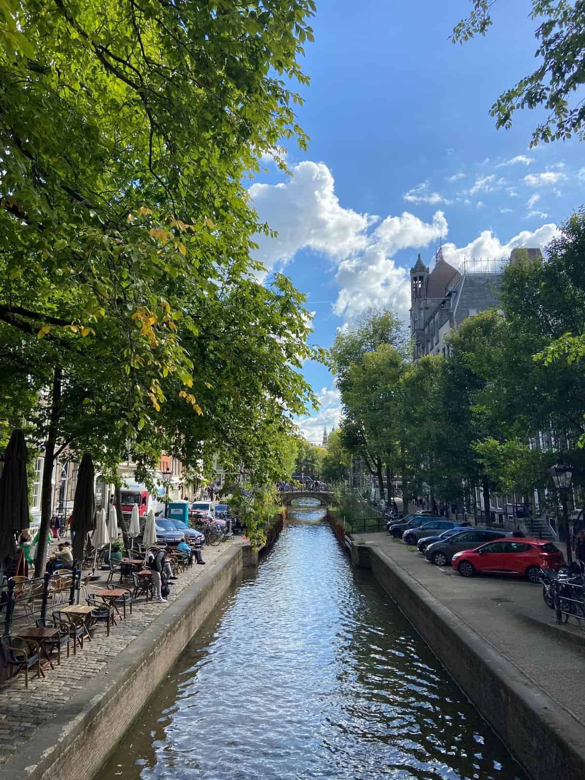 A view down a canal in Amsterdam flanked by trees, parked cars and cafe tables